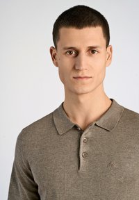 Polo marrón confeccionado en una tela suave, con cuello, tres botones y un sutil bordado del logo en el lado izquierdo del pecho.
