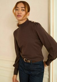 Brauner Strickpullover mit hohem Kragen, gewelltem Detail und Knopfakzenten an der Schulter. Kombiniert mit dunklen Jeans und einem braunen Gürtel.