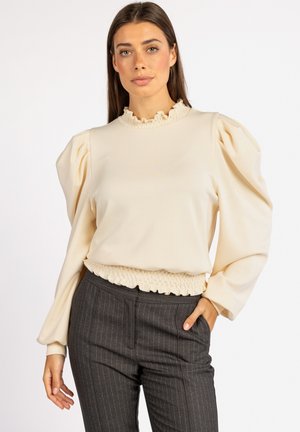 Crème-kleurige pullover met een ruches halslijn, volume mouwen en een gerimpelde tailleband. Gepaard met donkere gestreepte broek.