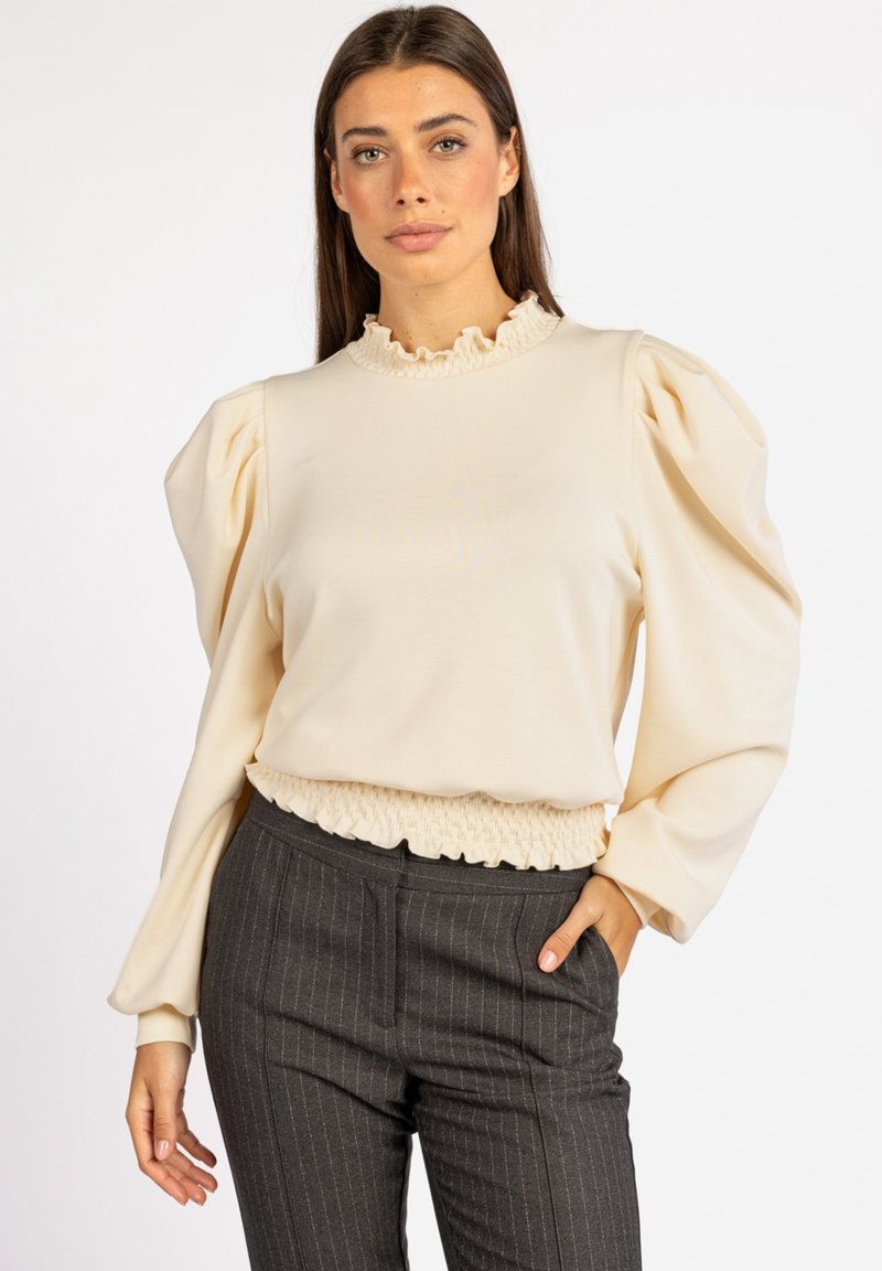 Crème-kleurige pullover met een ruches halslijn, volume mouwen en een gerimpelde tailleband. Gepaard met donkere gestreepte broek.