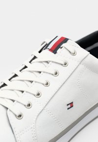 Vit sneaker med vita snören, silverfärgade öglor, liten röd, vit och blå logotyp på sidan, samt randig rem av röd, vit och svart tyg på tungan.