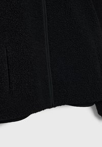 Gros plan d'une veste polaire noire texturée avec une couture verticale visible et une ouverture de poche latérale.