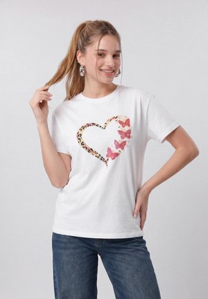 Νεαρή γυναίκα που χαμογελά, φορώντας λευκό T-shirt με σχέδιο καρδιάς από λεοπάρ εκτύπωση και ροζ πεταλούδες, συνδυασμένο με μπλε τζιν.