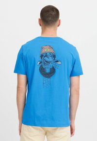 Modré bavlněné tričko s černou grafikou žirafy, která nosí hnědou čepici se štítkem "COOL", na jednoduchém pozadí.