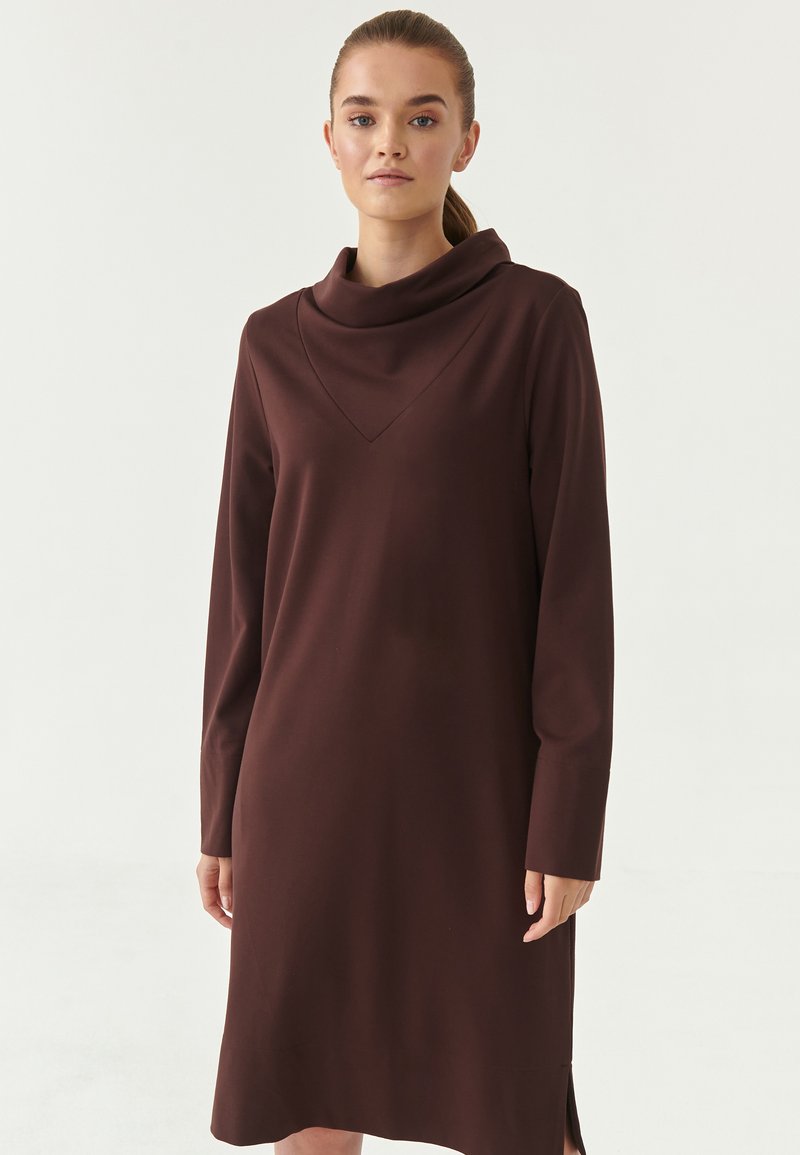 En brun knælængde kjole med en løs cowl-hals, lange ærmer og en trekantet detalje under halsen. Stof er glat med sideudskæringer.