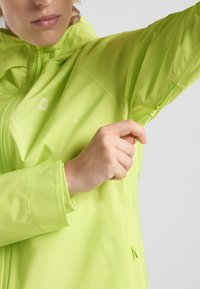 Helle limegrüne wasserdichte Jacke mit einem Reißverschluss vorne und verstellbaren B�ündchen. Verfügt über einen strukturierten Stoff und ein Logo auf der Brust.