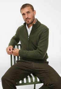 Donkergroene gebreide trui met een hoge kraag en ritssluiting, gedragen over een wit shirt, gecombineerd met donkere broeken, zittend op een groene bank.