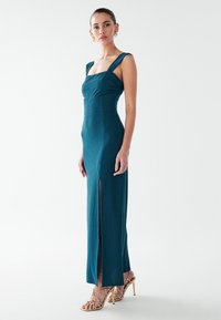 Teal tajt klänning med breda axelband, texturerat tyg och en slits högt upp på låret; kombinerad med guldiga skor med smala remmar och hög klack. Ren, minimalistisk bakgrund.