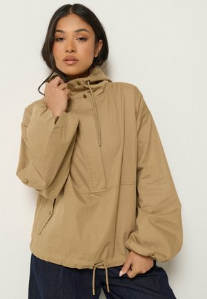Vrouw draagt een losse beige capuchonjas met koordjes, staand tegen een witte achtergrond, houdt met één hand de jaskraag vast.