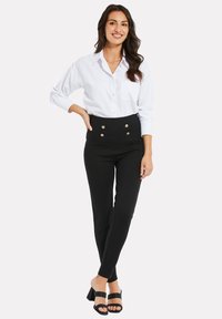 Witte lange mouwen shirt, gecombineerd met hoge zwarte broek met gouden knoopaccenten, afgewerkt met zwarte hoge hakken met brede band.