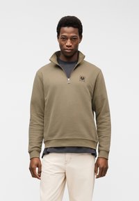 Junger Mann, der ein olivgrünes Sweatshirt mit halbem Reißverschluss über einem dunklen Hemd und cremefarbenen Hosen trägt, vor einem schlichten hellen Hintergrund steht.