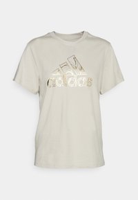 Beige bomulds-t-shirt med v-udskæring, der har et reflekterende guld Adidas-logo på tværs af brystet. Korte ærmer og en afslappet pasform.