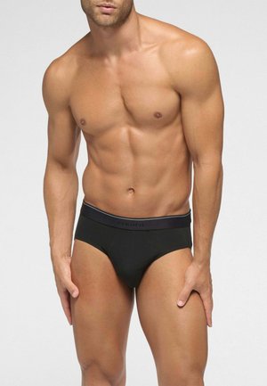 Uomo muscoloso che indossa slip neri aderenti con una fascia in vita firmata, in piedi davanti a uno sfondo chiaro e semplice.