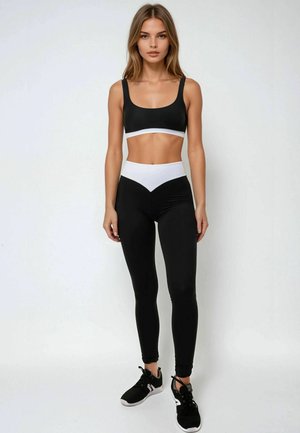 Zwarte sportbeha met witte band, gecombineerd met hoge zwarte leggings met een wit chevronpatroon, aangevuld met zwarte sportschoenen.