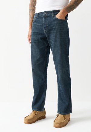 Mężczyzna ubrany w ciemnoniebieskie proste jeansy, białą koszulę i beżowe zamszowe buty sznurowane, z jedną ręką w kieszeni.