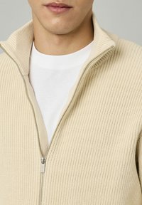 Un pull zippé côtelé beige avec un col haut, superposé à un t-shirt blanc. Il présente une fermeture éclair argentée et un tissu texturé.