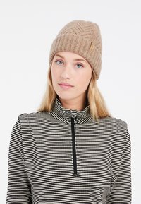 Beige Strickmütze mit strukturiertem Muster, versehen mit einem kleinen Lederetikett; das Model trägt ein schwarz-weiß gemustertes Zip-Up-Oberteil.