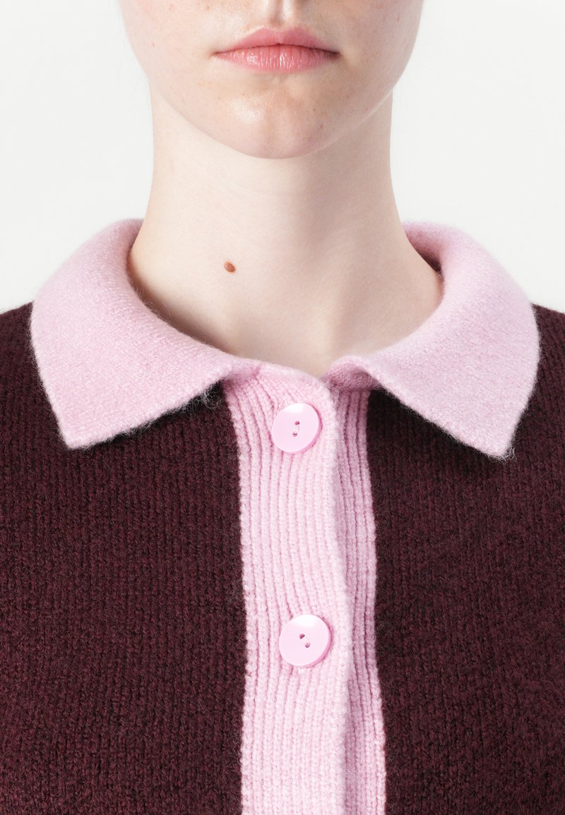 Bordeaux brei sweater met een roze kraag. Beschikt over een geribbelde middenknopenlijst en twee roze knopen. Zachte textuur met contrasterende kleuren.
