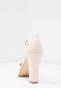 Beige högklackad sko med en lackläderfinish, strukturerad design och justerbart spänne runt ankeln.