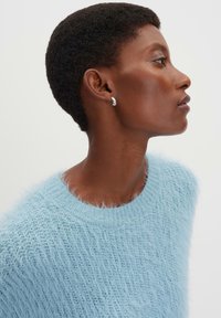 Hellblauer, fuzziger Strickpullover mit Rundhalsausschnitt und strukturierter Oberfläche, getragen von einem Model mit kurz geschnittenen Haaren und silbernen Ohrringen.