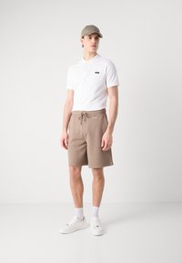 Biała koszulka polo z małym logo, beżowe szorty z troczkami i białe sneakersy. Strój charakteryzuje się prostym, casualowym designem i lekką teksturą.