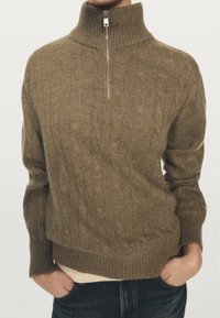Bruine kabelgebreide trui met hoge kraag en halve rits. Geribbelde manchetten en zoom, gedragen over een shirt in een lichtere kleur.