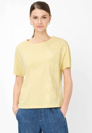 Frau mit braunen, zurückgebundenen Haaren, goldenen Creolen, einem hellgelben kurzärmeligen Hemd mit Blumenstickerei und blauer Jeans, die Hände in den Taschen.