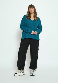 Turquoise knit sweater with a V-neck and side slits, paired with black cargo pants featuring pockets and elastic cuffs, worn with chunky sneakers.