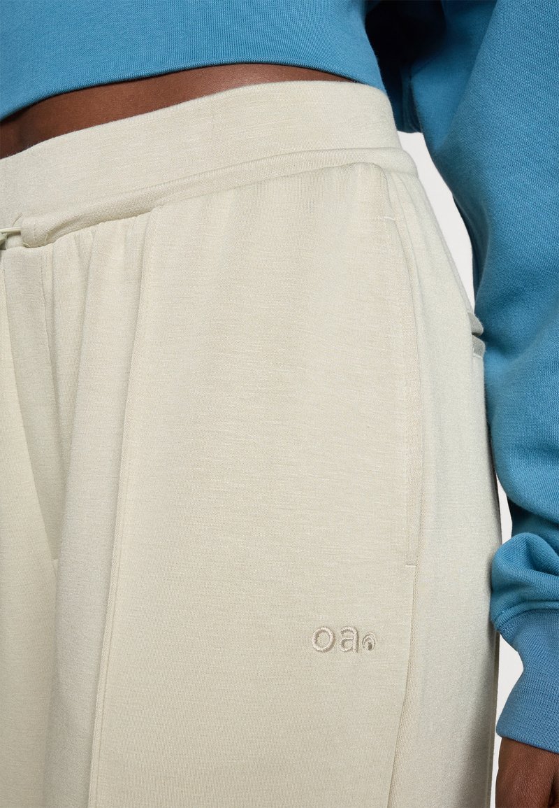 Pantaloni jogger beige chiaro con vita elastica, tasche laterali e piccolo logo ricamato in un tessuto morbido. Abbinati a una maglietta blu.