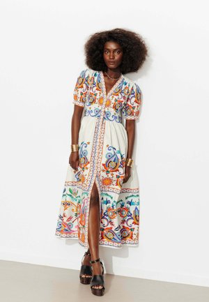 Femme aux cheveux bouclés portant une robe midi blanche à motif floral avec des manches bouffantes et une fente avant, debout contre un mur blanc uni.