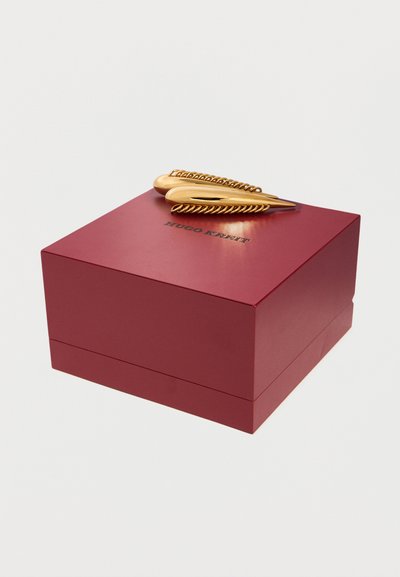 Βαθύ κόκκινο κουτί Hugo Kreft με δύο λαμπερά χρυσά διακοσμητικά κλιπ μαλλιών τοποθετημένα από πάνω σε απλό λευκό φόντο.