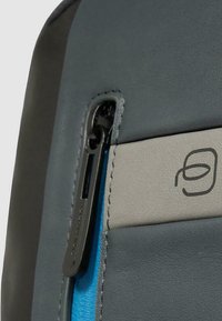 Bolsa de cuero gris con detalle de cremallera azul. Presenta una tira de acento gris y una lengüeta de cremallera negra con un logo. Superficie texturizada en toda la pieza.