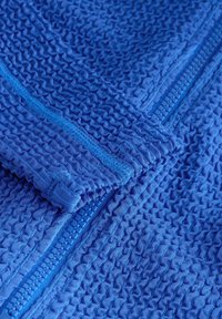 Mėlynos spalvos tekstūruota medžiaga su bangų raštu ir užtrauktuku. Medžiaga atrodo lengva ir lanksti, tinkama kasdieniam dėvėjimui arba aktyviam naudojimui.