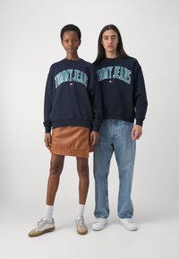 Sudaderas de cuello redondo en color marino con el texto "TOMMY JEANS", combinadas con una falda de cuero marrón y jeans de color azul claro, llevadas con zapatillas deportivas.