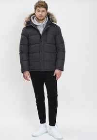 Chaqueta de plumas negra con capucha forrada de piel, que presenta un cierre frontal con cremallera y botones, combinada con pantalones negros y zapatillas blancas.
