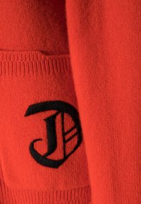 Czerwony wełniany sweter z wyraźnym czarnym haftowanym logo. Tkanina ma miękką strukturę z prążkowanymi detalami na dole i kieszeni.