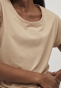 Beige bomullströja med rund halsringning och korta ärmar. Mjuk textur och enkel design, bärs med armarna korsade.