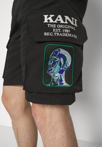 Svarte cargo-shorts i teksturert stoff. Har hvit brodert logo over et fargerikt, detaljert portrettlapp på låret.