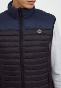 Schwarw und marineblau gepolsterte Weste mit hohem Kragen, Frontreißverschluss und einem runden Logopatch auf der Brust. Weicher, gestepptes Material.