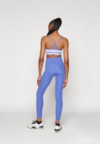 Blauwe sportbeha met kruislingse banden en hoge blauwe legging. Schoenen zijn wit met zwarte en gele accenten. Glad, rekbaar stof.