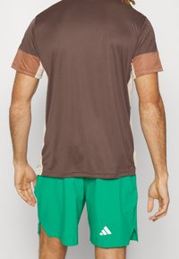 Camiseta deportiva marrón con acentos beige, combinada con pantalones cortos verdes que tienen un logo blanco. Tela suave, mangas cortas y corte relajado.