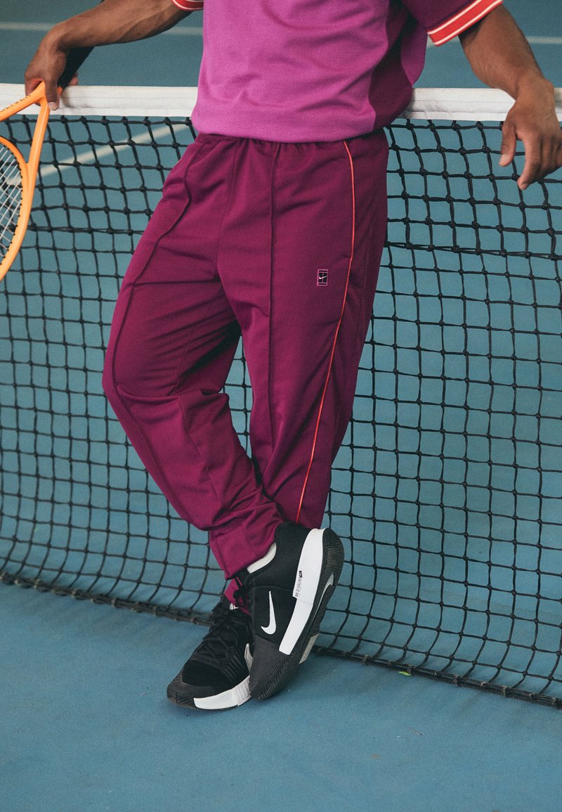 Pantaloni sportivi viola con strisce laterali arancioni, abbinati a scarpe Nike nere. Una racchetta da tennis è tenuta vicino a una rete su un campo blu.