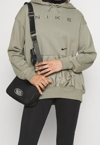 Olivově zelená mikina Nike s černým logem, s texturovanou přední kapsou. V kombinaci s černými legínami a malou černou kabelkou přes rameno.