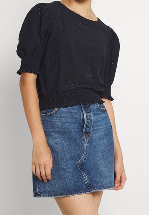 Vrouw draagt een zwarte blouse met pofmouwen van eyelet-stof, een getailleerde taille met smockwerk en een korte blauwe denim rok.