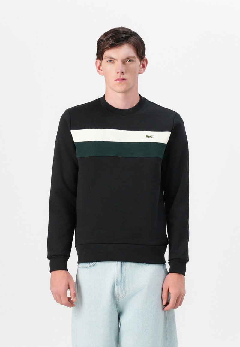 Svart hettegenser med en horisontal hvit og grønn stripe over brystet; har rund hals og viser logo. Myk stoff.