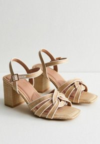 Beige woven heeled sandals feature a square toe, crisscross straps, and a block heel with a textured design. Adjustable ankle strap included.
