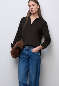 Blusa negra de manga larga con cuello, combinada con jeans de pierna ancha azules. La modelo sostiene un bolso de mano de ante marrón. Fondo neutro.