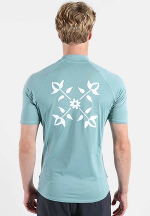 Homme aux cheveux blonds courts portant une chemise bleu clair à manches courtes avec un motif floral blanc dans le dos, debout devant un fond uni.