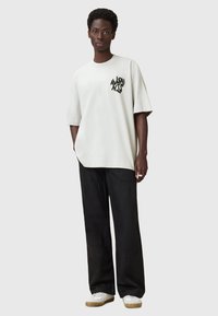 Joven con cabello rizado corto que lleva una camiseta blanca oversized con texto negro, pantalones negros holgados y zapatillas blancas, de pie contra un fondo liso.