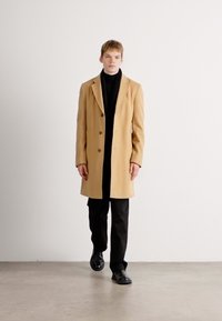 Beige wool coat with a tailored design, three large buttons, and a notched collar, worn over a black turtleneck and black trousers.